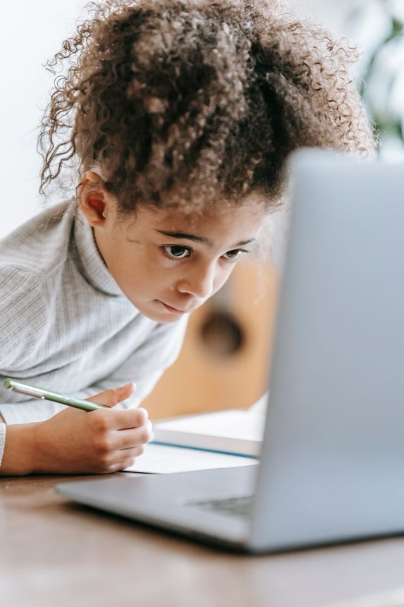 A young student concentrating on an online lesson at home, writing beside her laptop